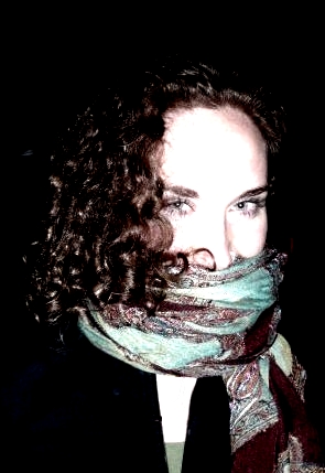 Julia Voss headshot, an over-exposed close-up against a dark backdrop of my face (pale skin, brown curly hair) with a green and purple scarf wrapped around the lower half of my face, obscuring it